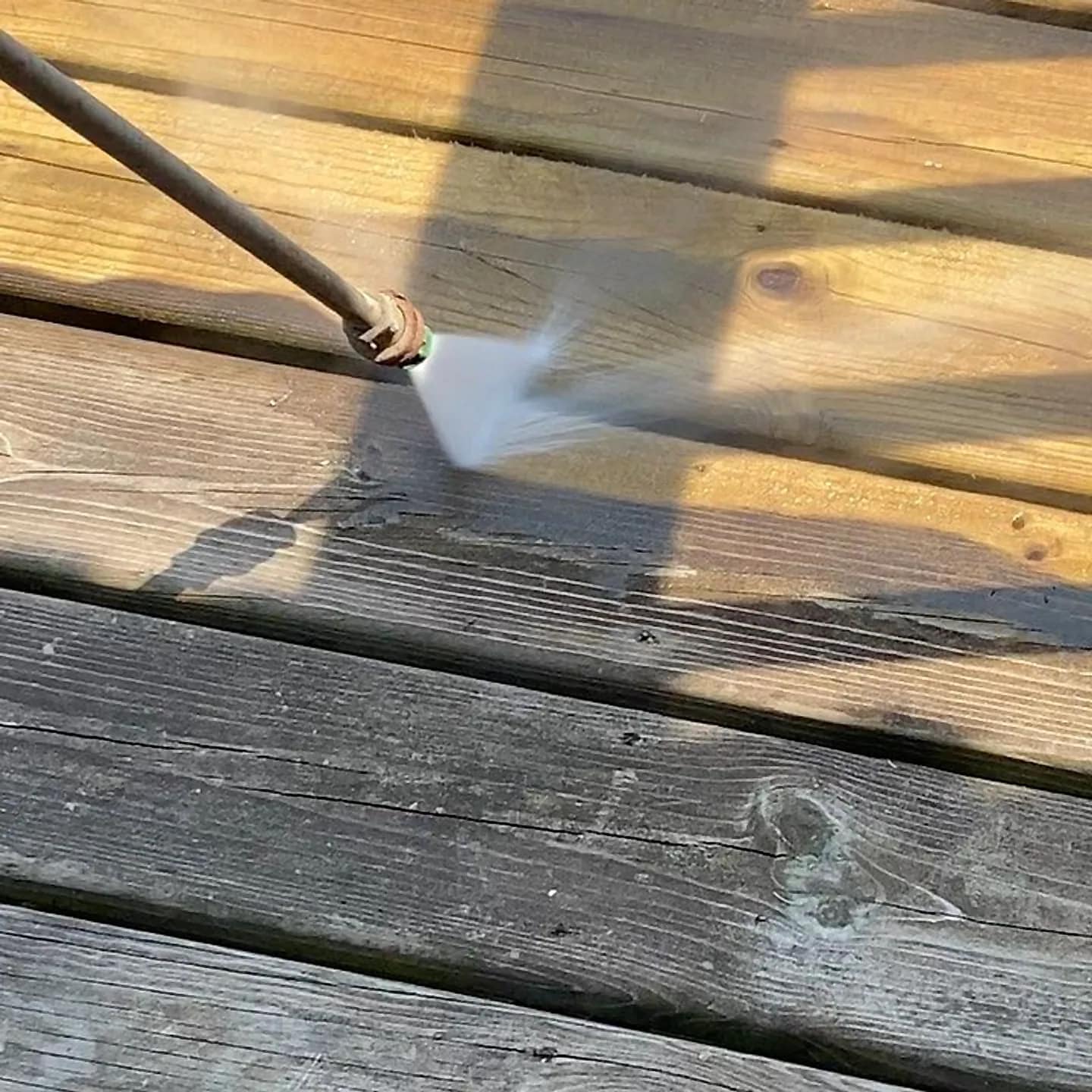 Wood deck being pressure washed, the wand spraying a bright gold-clean stripe through weathered grey boards